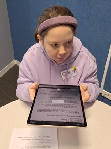 Figure 2 Chevarni stands beside a table, holding an iPad with the Seeing AI app positioned above a printed recipe document which is formatted in Microsoft Word. She is wearing a purple sweatshirt and a purple headband.