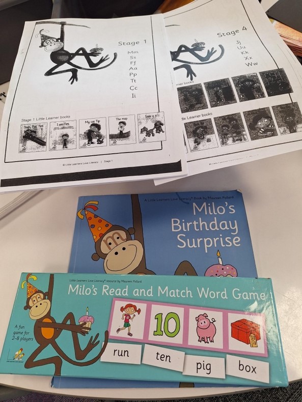Four examples of structured literacy resources sitting on a table. There is two worksheets, one reading book and one game.