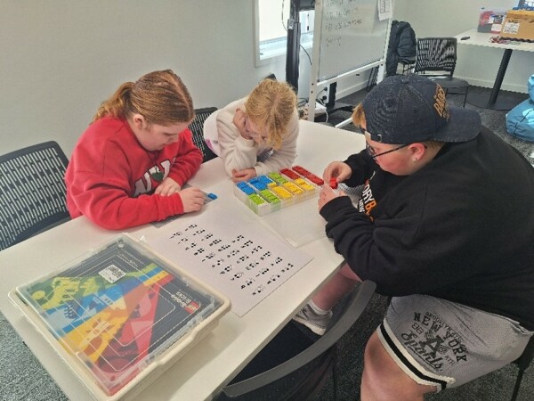 Three ākonga are sitting on chairs at a table exploring a set of Braille bricks. The boy is sitting on one side of the table and has his cap on backwards. There is two girls sitting on the other side. They both have their hair pulled back onto ponytails.