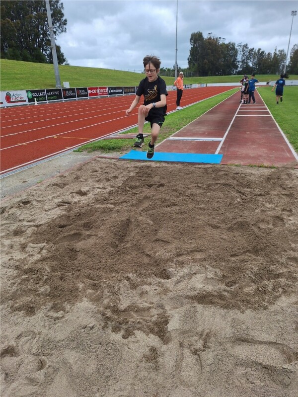 Ākonga is mid air after taking off from long jump launch pad and is about to land in the sand. There is an adult and a group of ākonga standing in the background.