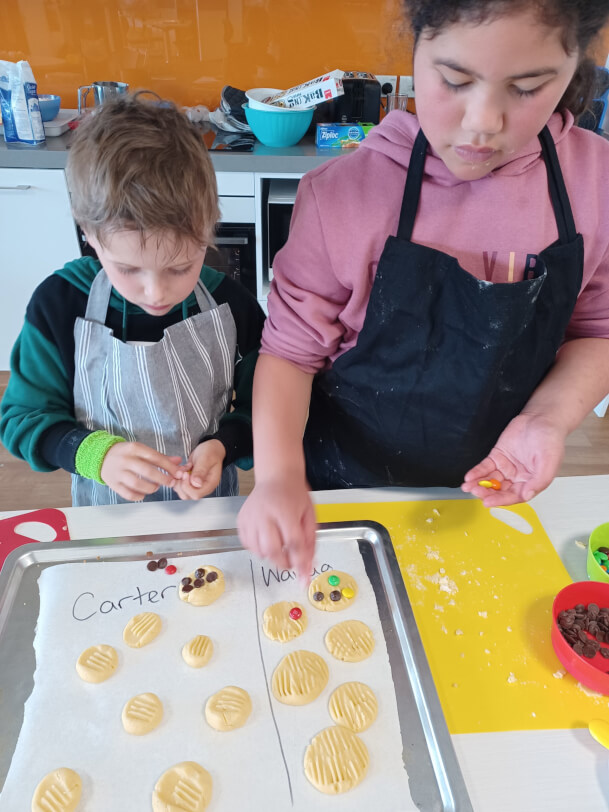 A boy and girl wearing aprons place decorations onto cookies on a tray.