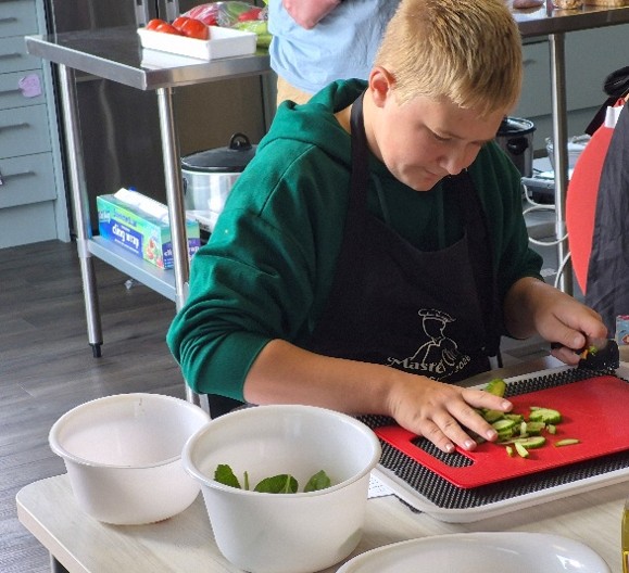 Figure 1 A young person in a green hoodie and black apron is slicing cucumbers on a red cutting board in a kitchen setting