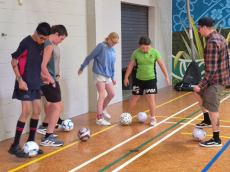 Five people in a gymnasium practicing soccer ball control, each with a soccer ball at their feet. There are two boys (Ranveer and Kahn) and two girls (Auti and Charlene) and one adult (Stuart).  They are all wearing casual sportswear, positioned along a wall with gym floor markings visible